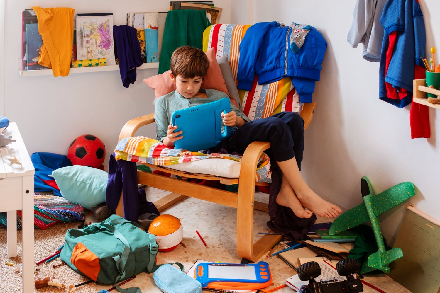 A boy sits barefoot in a colorful, cluttered room on a chair, using a tablet surrounded by scattered toys, clothes, and school supplies.
