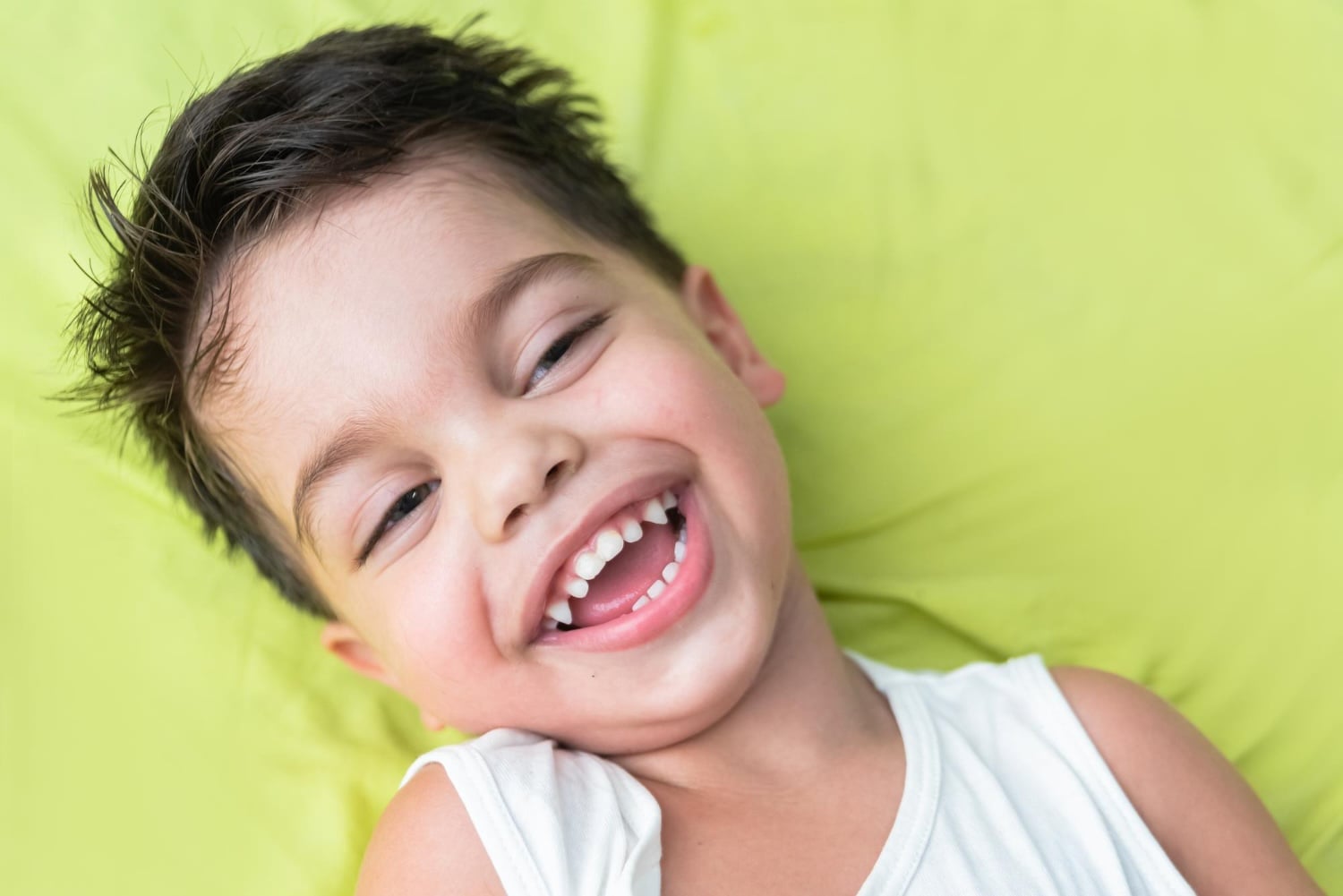 A young boy with dark hair smiles widely while lying on a bright green surface, wearing a white sleeveless shirt.