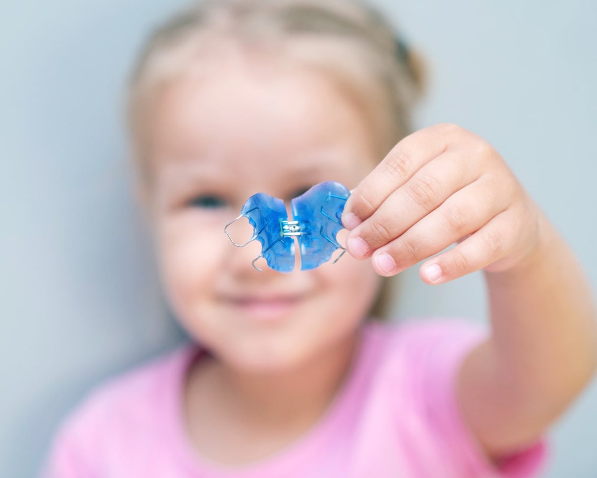 A young girl in a pink shirt holds a blue dental retainer in front of her, with her face blurred in the background.