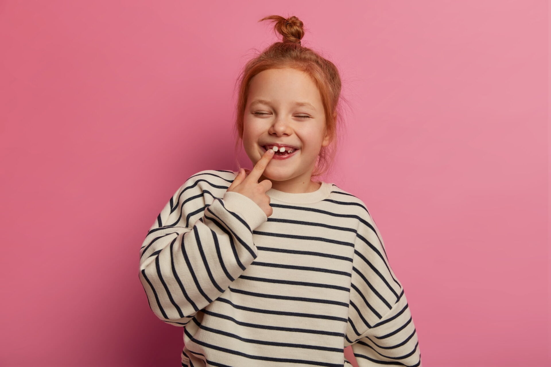 Child with red hair in a striped sweater stands in front of a pink background, smiling with eyes closed and pointing to a gap in their teeth.