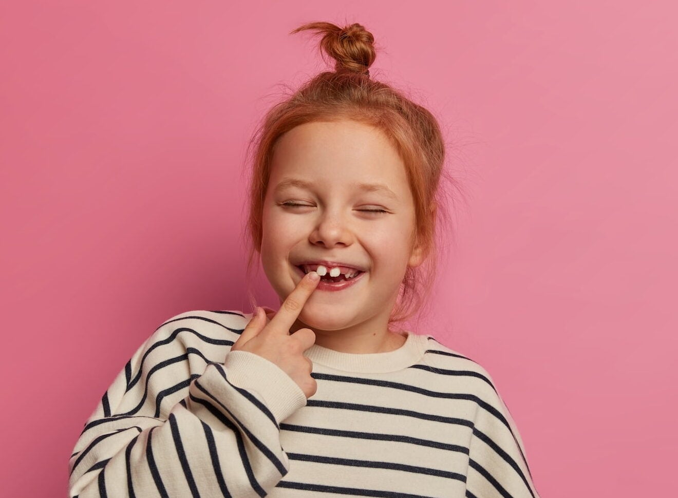 Child with red hair in a striped sweater stands in front of a pink background, smiling with eyes closed and pointing to a gap in their teeth.