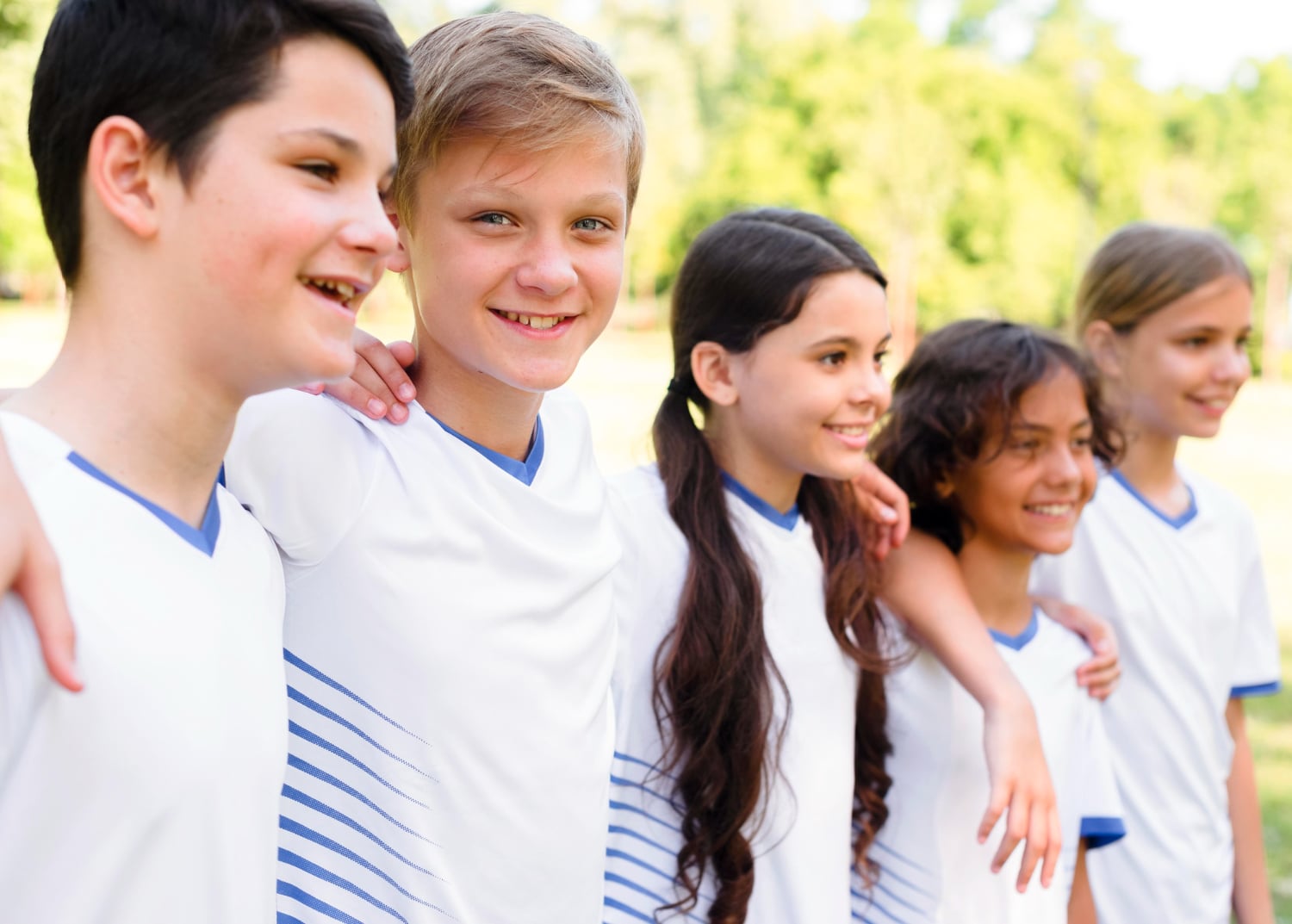 Five children wearing white sports jerseys stand in a row outdoors, smiling with arms around each other’s shoulders. Trees and greenery are visible in the background.