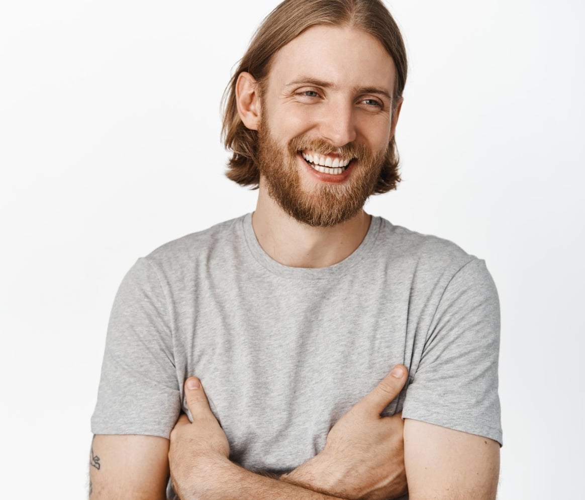 A man with shoulder-length brown hair and a beard smiles while standing with his arms crossed, wearing a light gray t-shirt against a plain white background.