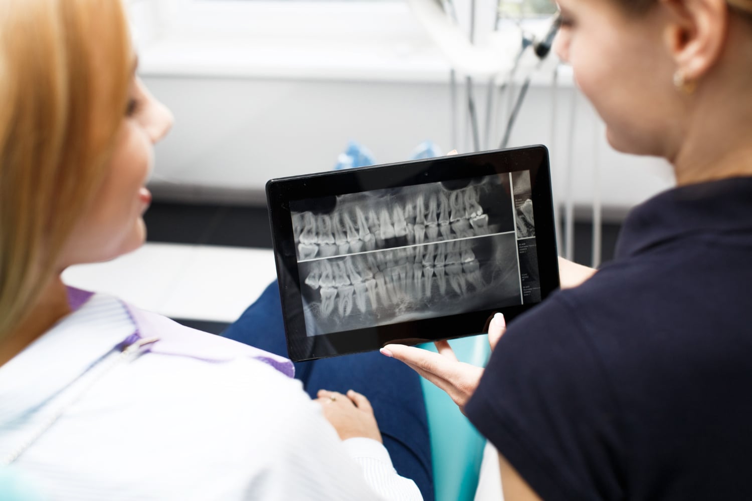 A dental professional shows a patient a digital X-ray image of teeth on a tablet during a dental consultation.