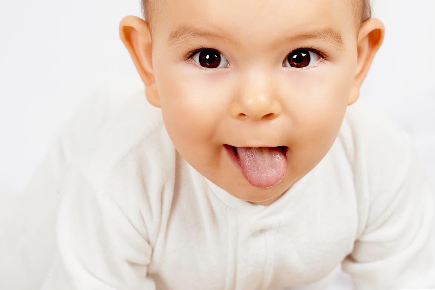 A baby in a white outfit sticks out their tongue, looking up at the camera against a plain light background.