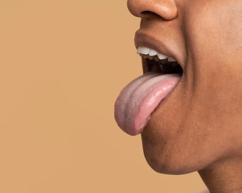 Close-up of a person with medium brown skin sticking out their tongue, mouth open, against a plain beige background.