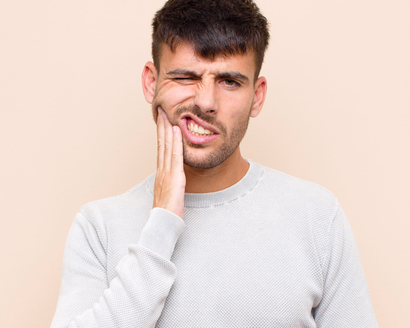 A man in a light gray sweater touches his cheek and winces, suggesting discomfort or pain, against a plain beige background.
